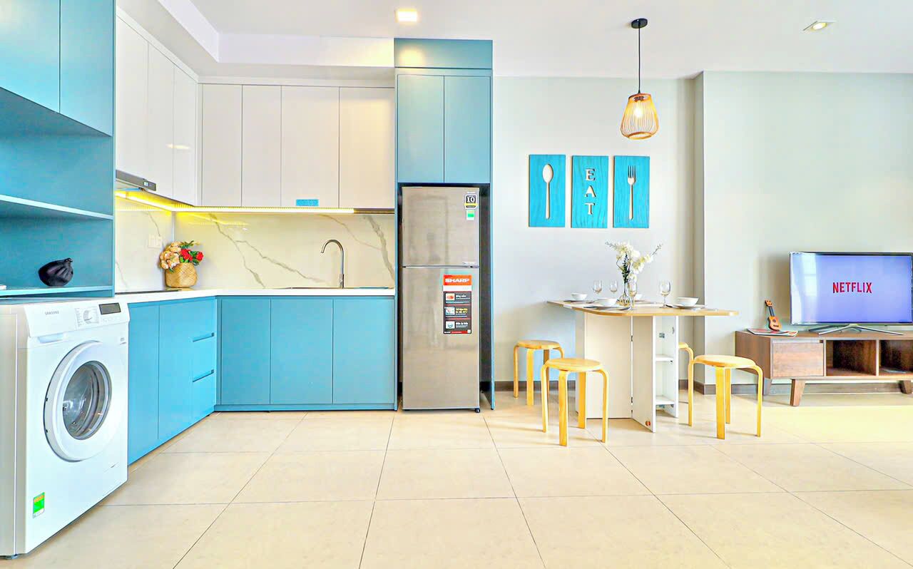 A modern kitchen with blue cabinets, a washing machine, a refrigerator, and a dining area with stools.
