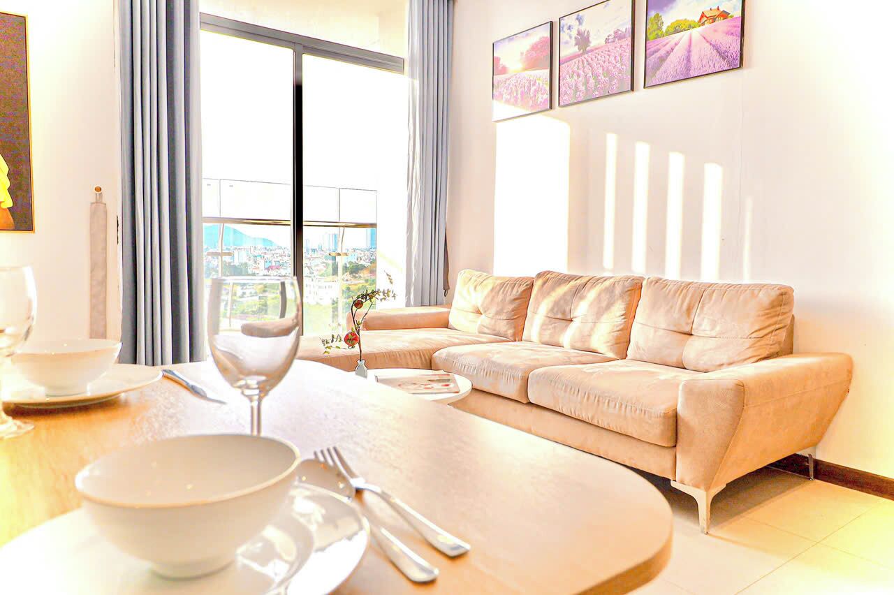 Een moderne woonkamer met een beige bank, een eettafel met een schaal, borden en bestek, en een groot raam met uitzicht op de stad.
