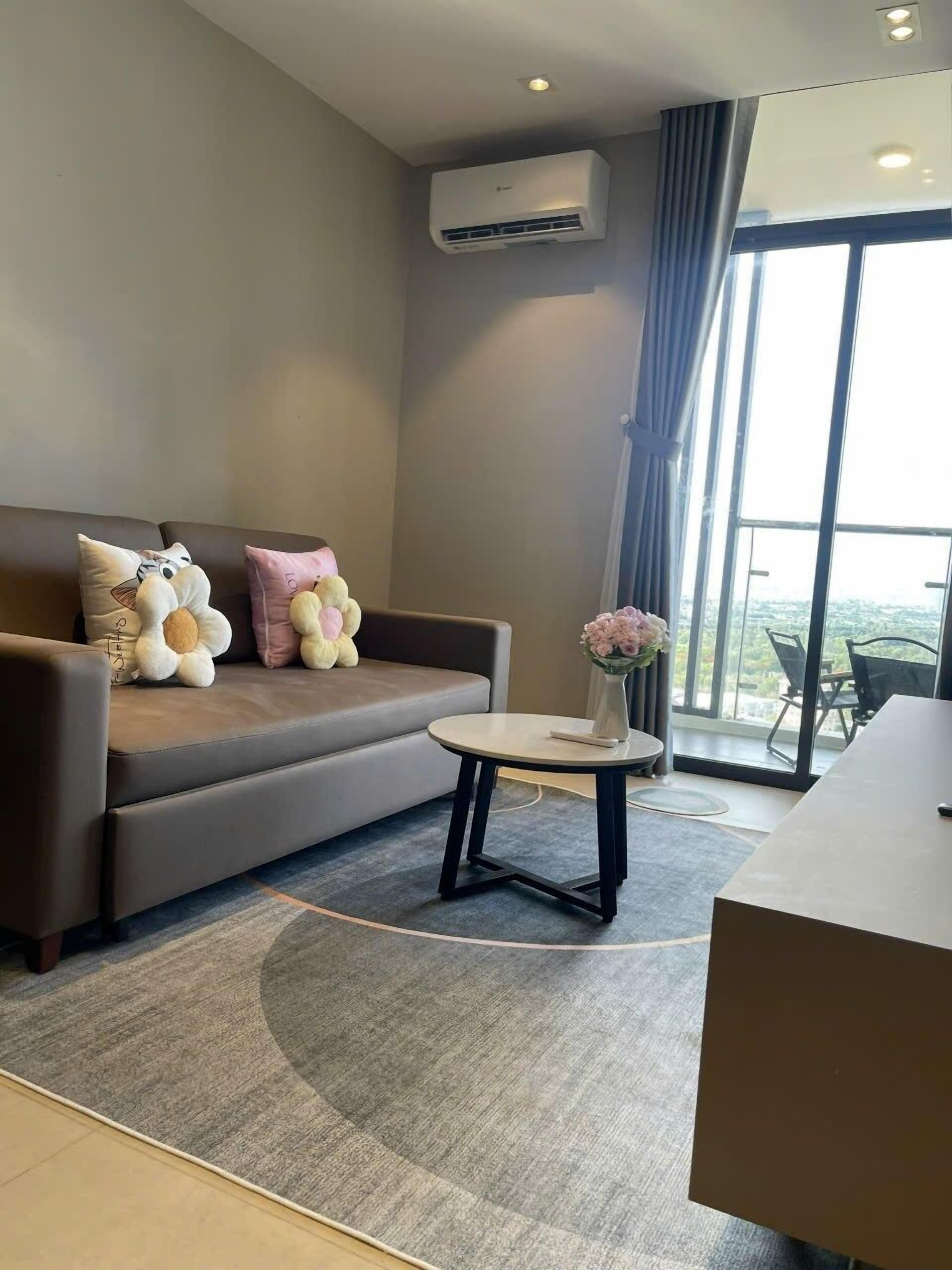 A modern hotel room with a grey sofa, a small round table, and a large window offering a view of the city.