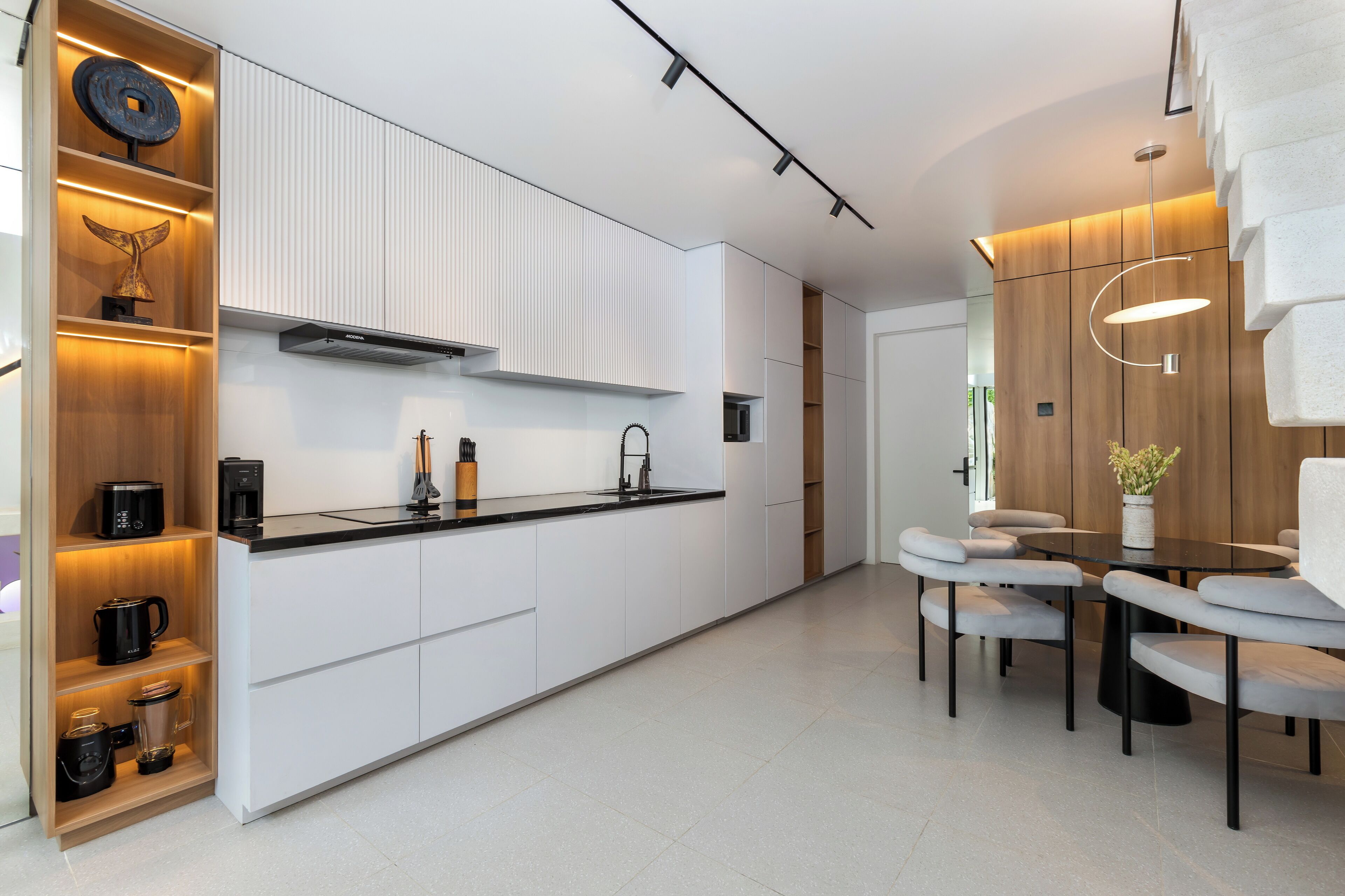Une cuisine moderne avec des meubles blancs, un plan de travail noir et un appareil à café intégré. On y trouve également un coin repas avec une table ronde et des chaises à revêtement gris.