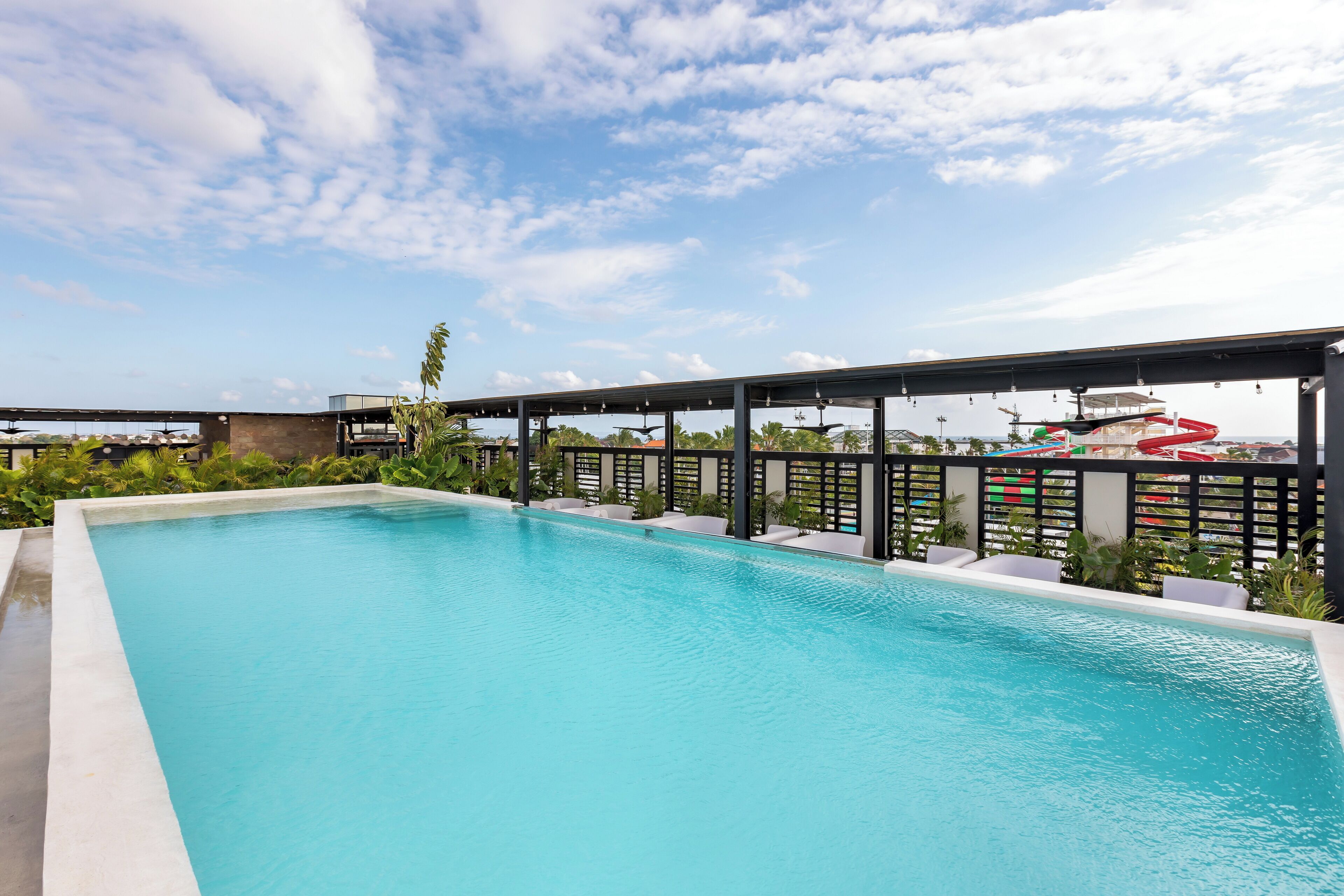 Uma piscina retangular com superfície de água azul translúcida, rodeada por uma área de deck coberta com assentos e plantas em vasos.