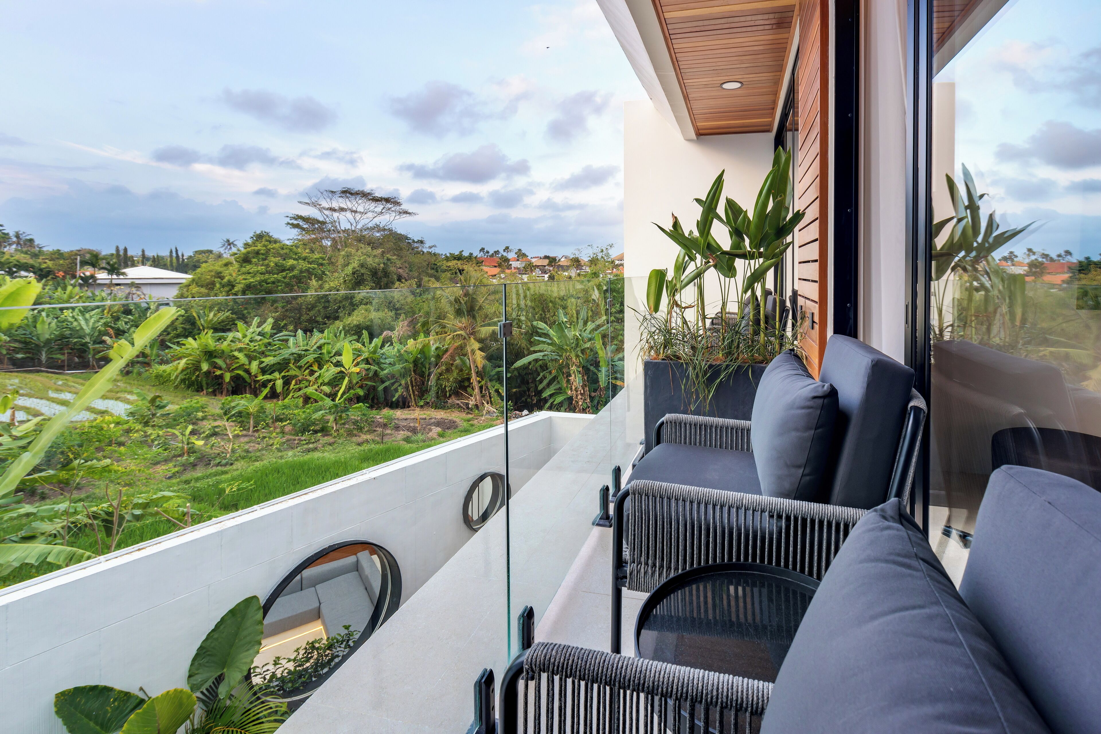 A balcony with modern wicker furniture, a glass railing, and a view of a lush garden and distant buildings.