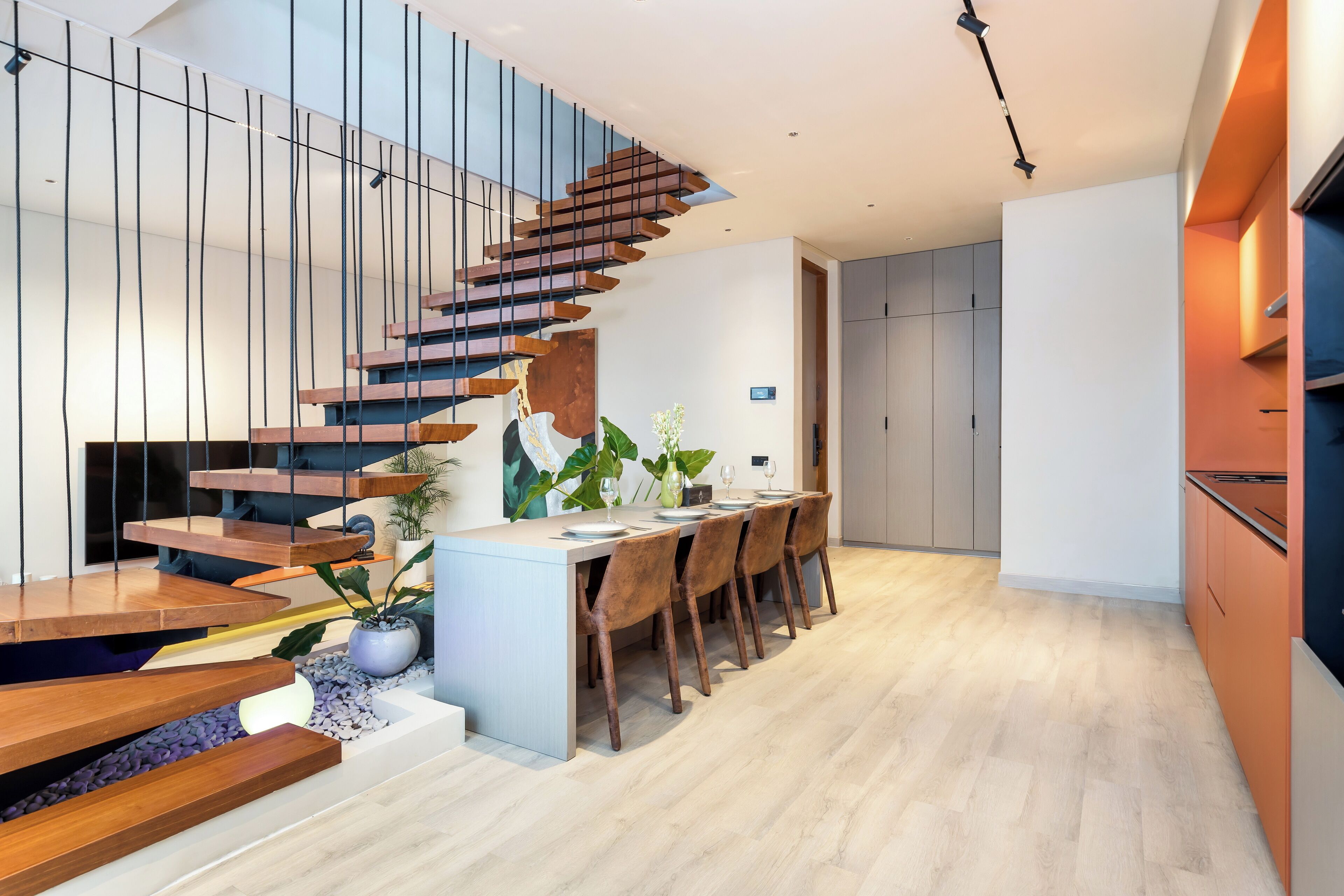 A modern kitchen with wooden cabinetry, a dining area with a long table and chairs, and a staircase with wooden steps.