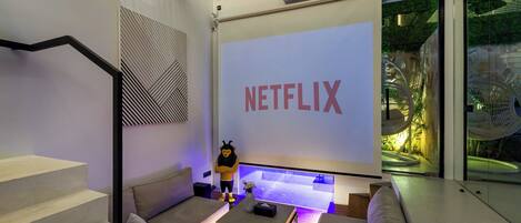 Una sala de estar moderna con una pantalla grande mostrando "NETFLIX", un sofá, una mesa de centro y una escalera.