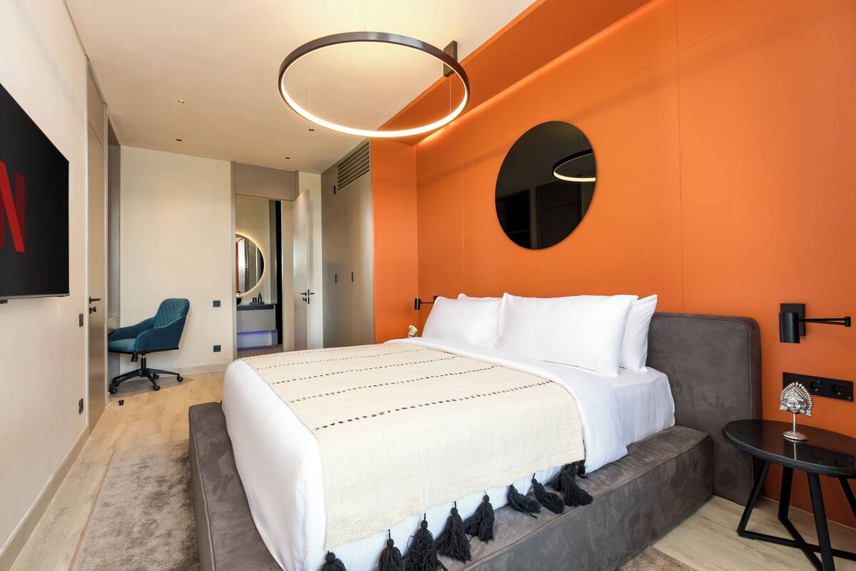 A modern hotel room with a large bed, a round mirror, a small table, and an orange accent wall.