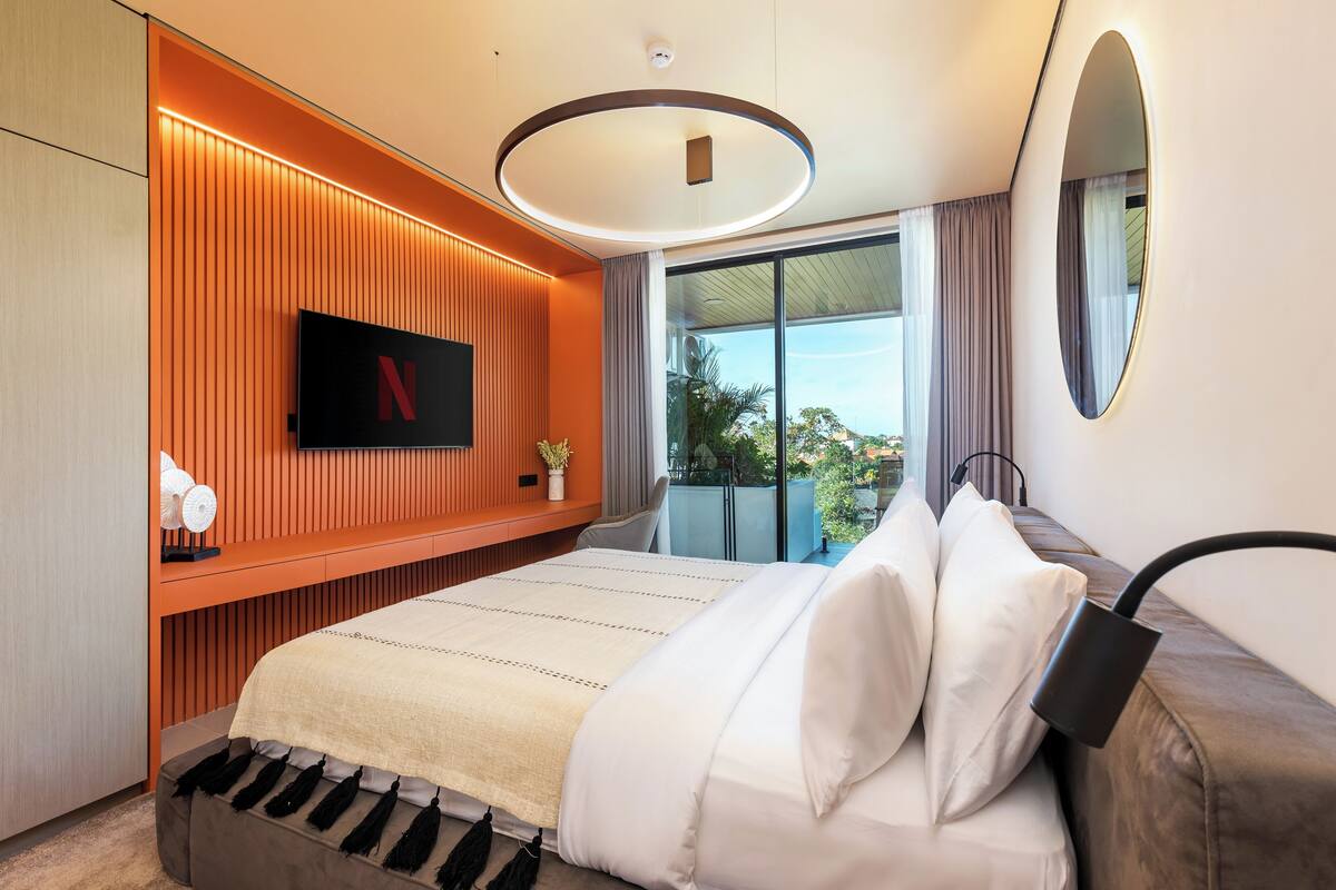 A modern hotel room with a large bed, a flat-screen TV mounted on the wall, a balcony with a view, and a circular ceiling light.