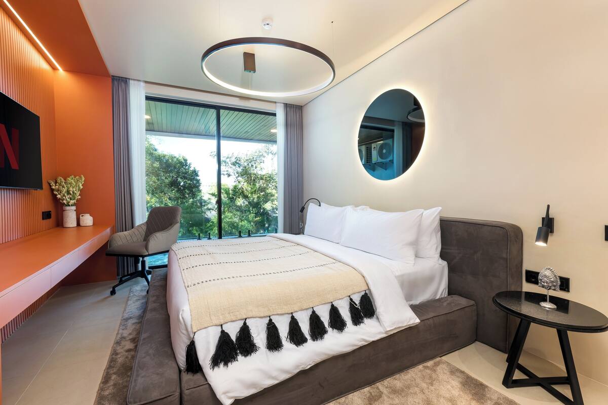 A modern bedroom with a large bed, a built-in desk, a round mirror, and a view of greenery outside.