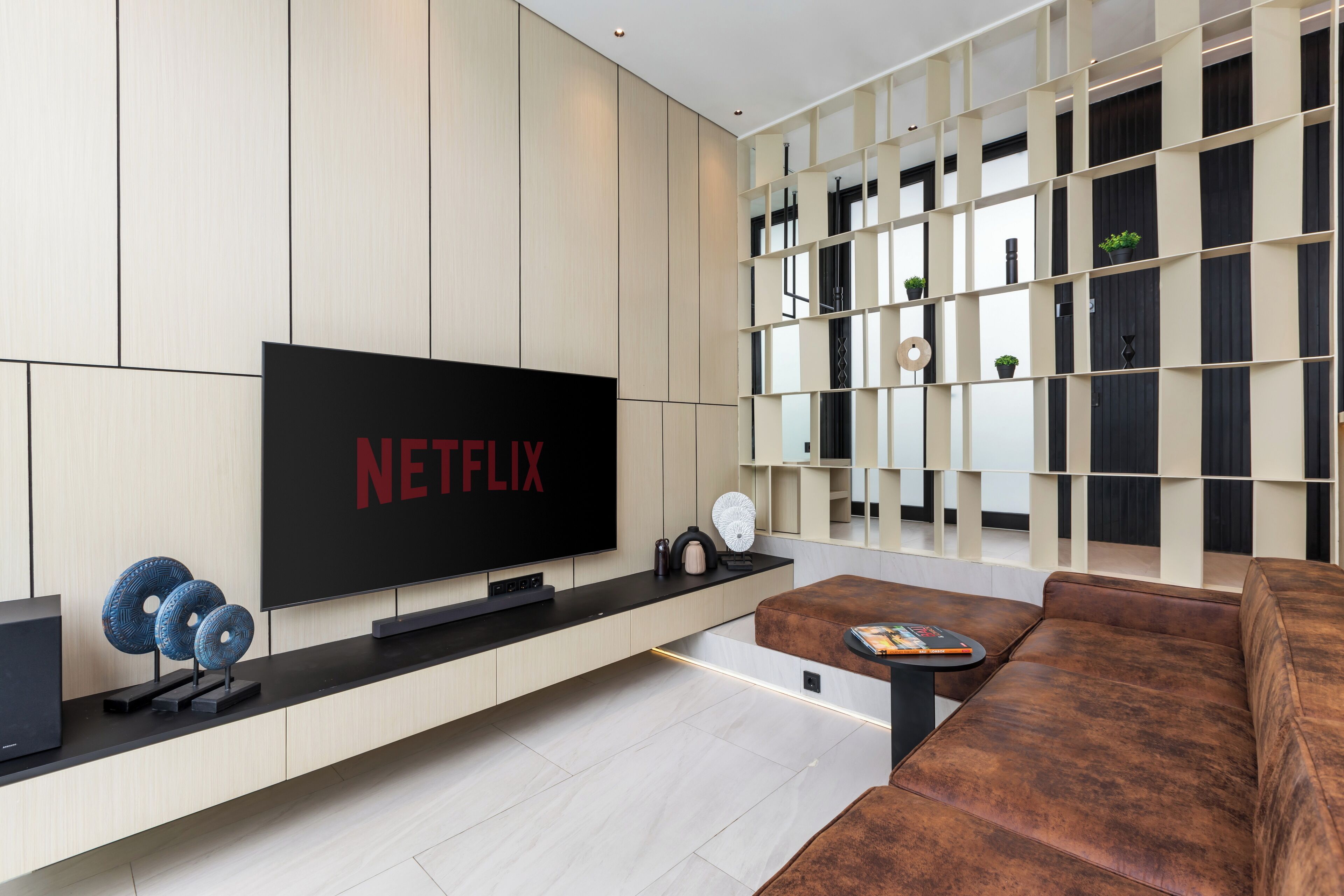 Un soggiorno moderno con una grande televisione a schermo piatto che mostra “NETFLIX”, un tavolino da caffè nero, un divano in pelle marrone e una parete attrezzata con oggetti decorativi.