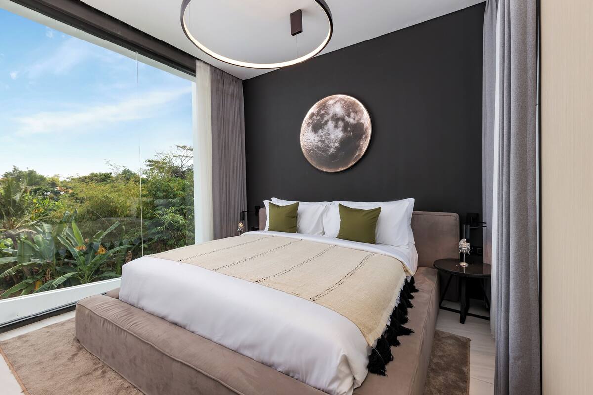 A modern bedroom with a large bed, a round wall art piece, and a view of greenery outside.