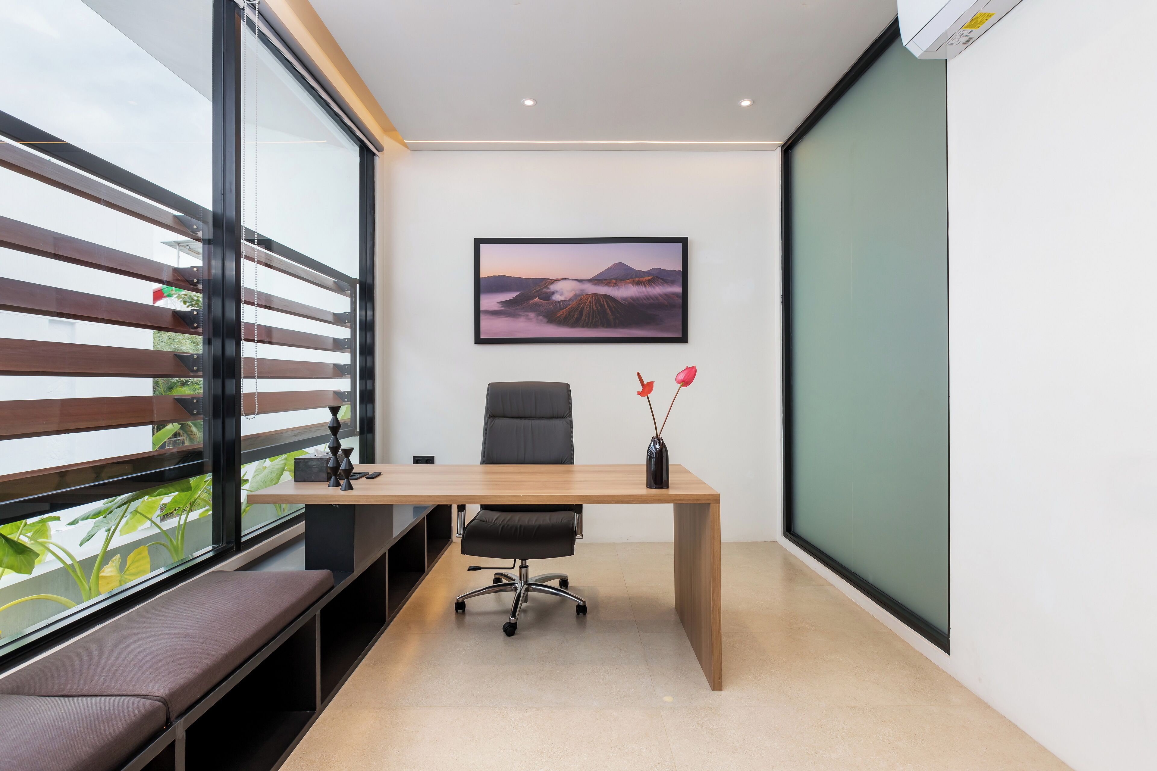 A modern office space with a desk, chair, and bench by a large window.
