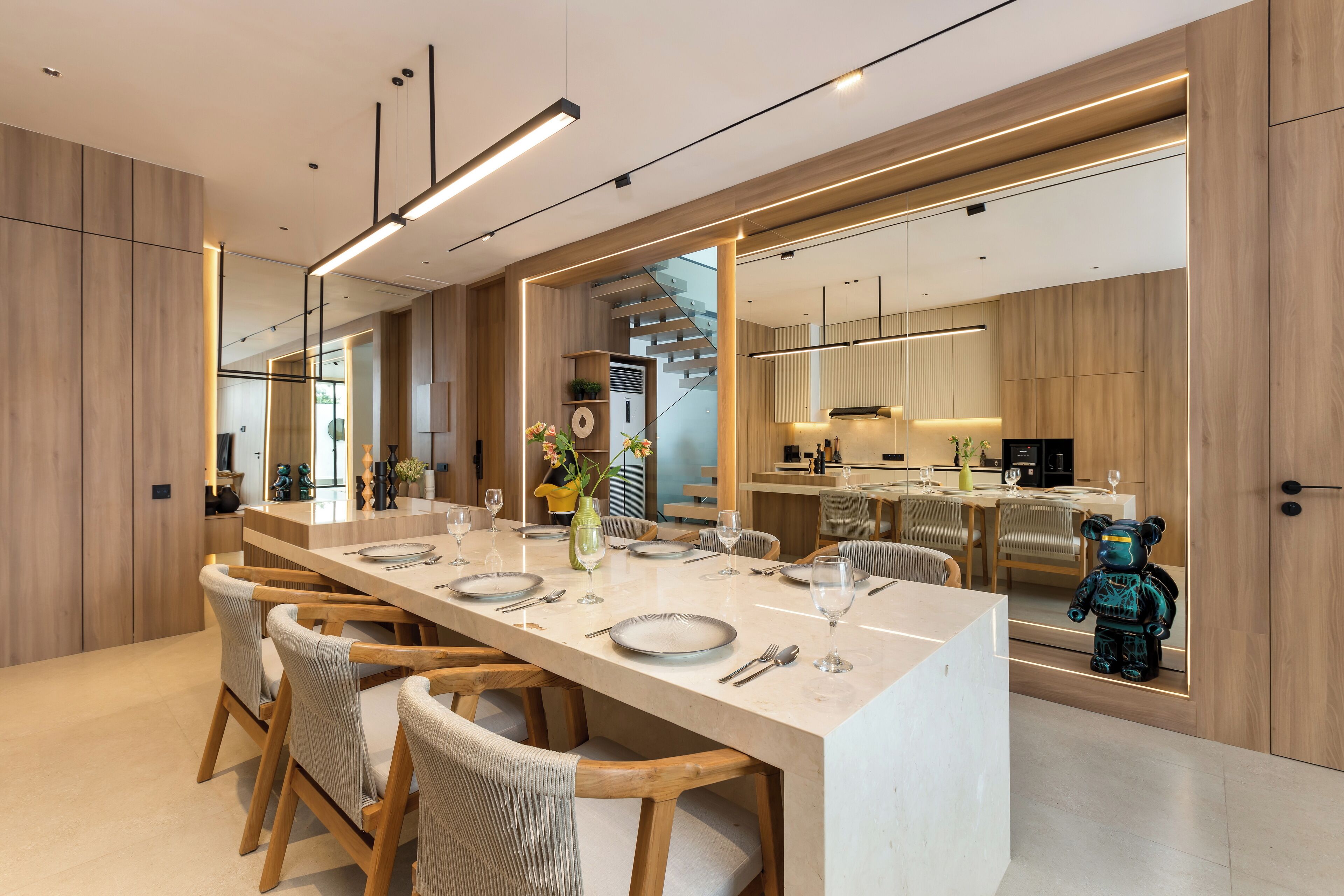 A modern dining area with a long table, wooden chairs, and a kitchen in the background.