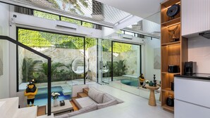 A modern interior with a glass-enclosed pool, a seating area, and a kitchen with wooden shelves. - Alex Villas Complex N5 (Canggu)
