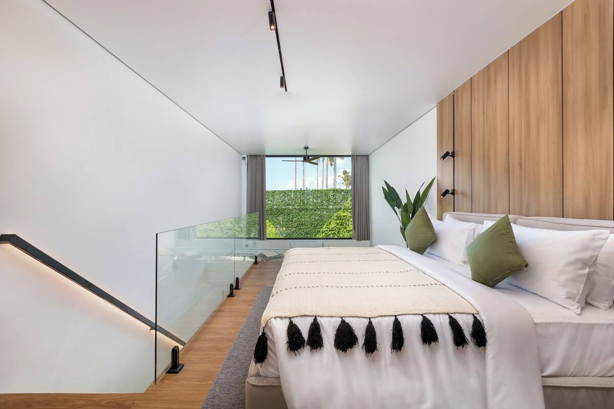 A modern bedroom with a large bed, a glass railing, and a view of greenery outside.