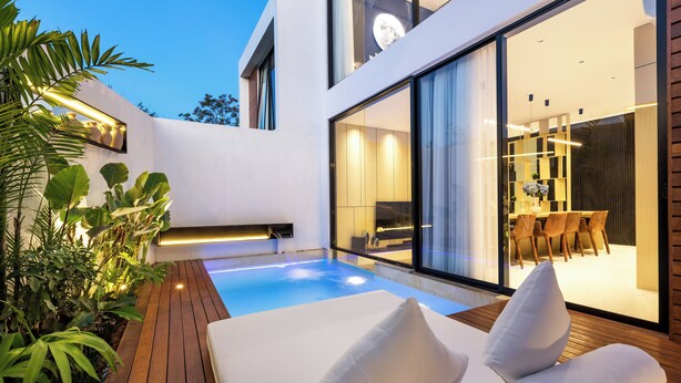 A modern outdoor pool area with a lounge chair and cushions, surrounded by lush greenery and a dining area visible through large glass doors. - Alex Villas Complex N5 (Canggu)