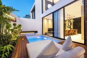 A modern outdoor pool area with a lounge chair and cushions, surrounded by lush greenery and a dining area visible through large glass doors. - Alex Villas Complex N5 (Canggu)