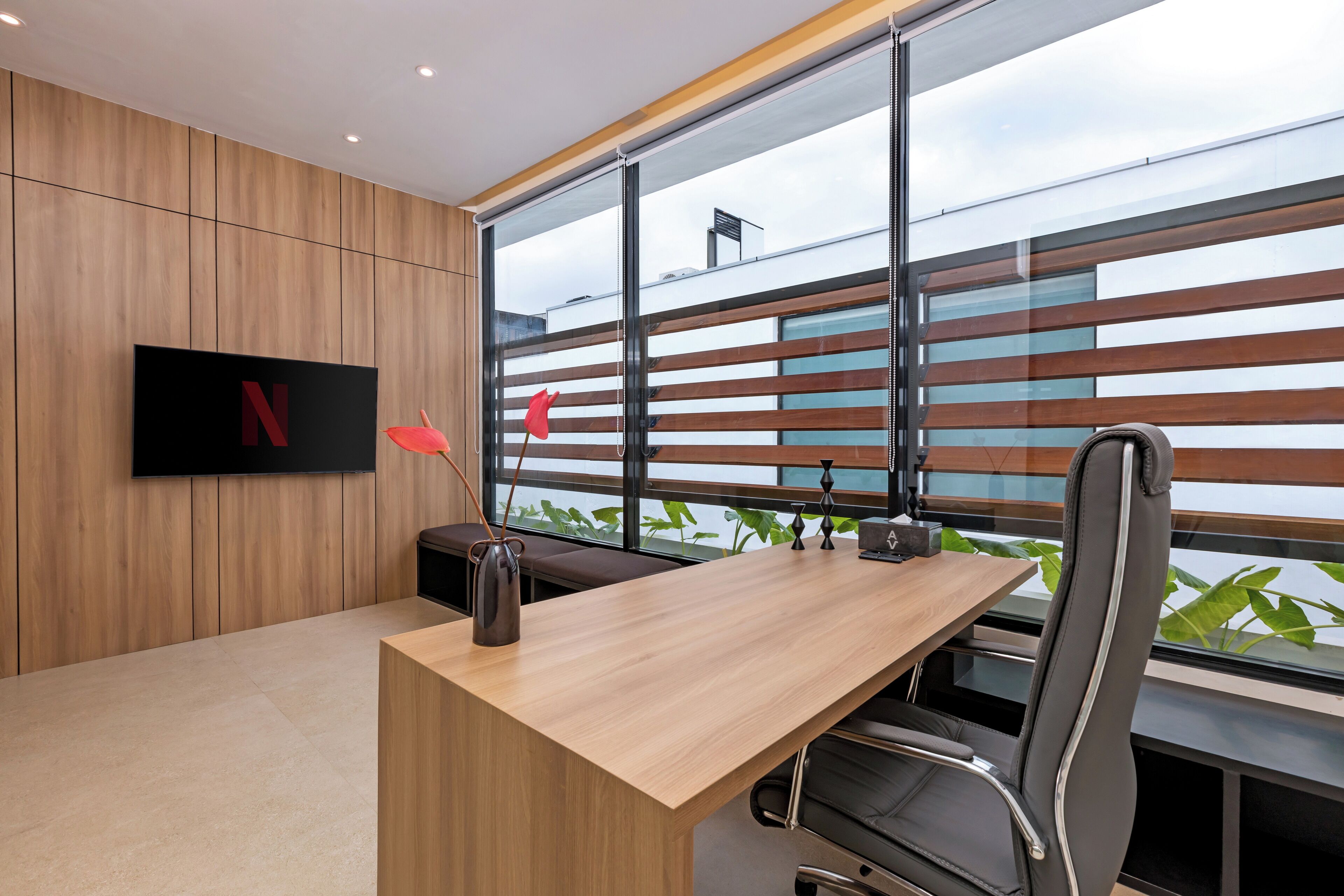 A modern office space with a wooden panel wall, a large window with blinds, a desk with a vase, and a chair.