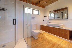 A modern bathroom with a glass-enclosed shower, a white toilet, and a wooden vanity with a large mirror. - Ocean Belle Onetangi (Waiheke Island)