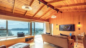 Amplia sala de estar con un gran ventanal que ofrece vista al mar, un sofá cómodo, un televisor y un techo de madera.
