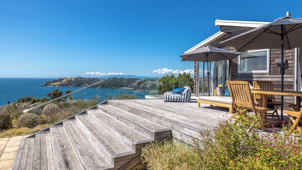 Una terraza de madera con mobiliario exterior, una barandilla de cristal y vista al océano.