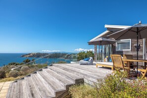 Eine Holzterrasse mit Gartenmöbeln, einer Glasreling und Blick auf den Ozean.