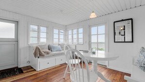 Ein Zimmer mit weißen Holzwänden und -decken, großen Fenstern, einem weißen Tisch mit Stühlen und einer gemütlichen Sitzecke mit Kissen.