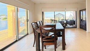 A spacious dining area with a large table and chairs, large windows with a view of a waterfront, and a balcony with outdoor furniture. - StayAU 5BR Lakefront Escape Point Cook (Point Cook)