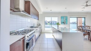 A modern kitchen with a large island, stainless steel appliances, and a view of a body of water through large windows. - StayAU 5BR Lakefront Escape Point Cook (Point Cook)