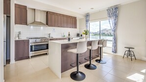 A modern kitchen with a central island, bar stools, and a view of the outside through large windows. - StayAU 5BR Lakefront Escape Point Cook (Point Cook)