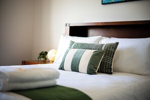 A neatly made bed with white and green bedding, a wooden headboard, and a bedside table with a lamp and a small plant.