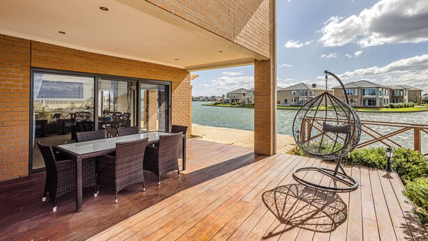 A wooden deck with a table and chairs, a hanging chair, and a view of houses across a body of water. - StayAU 5BR Lakefront Escape Point Cook (Point Cook)
