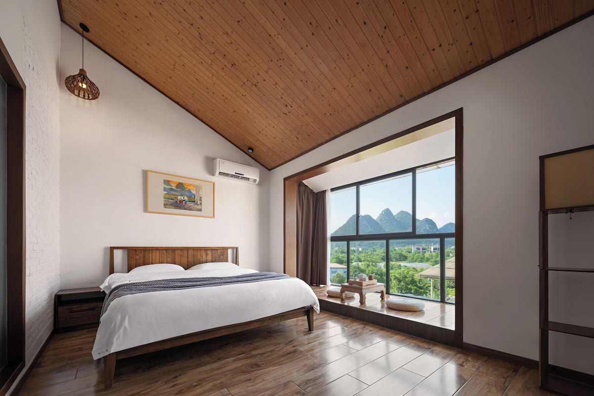 A bedroom with a wooden ceiling, a large window with a mountain view, a bed with a wooden headboard, and a small table with a lamp.