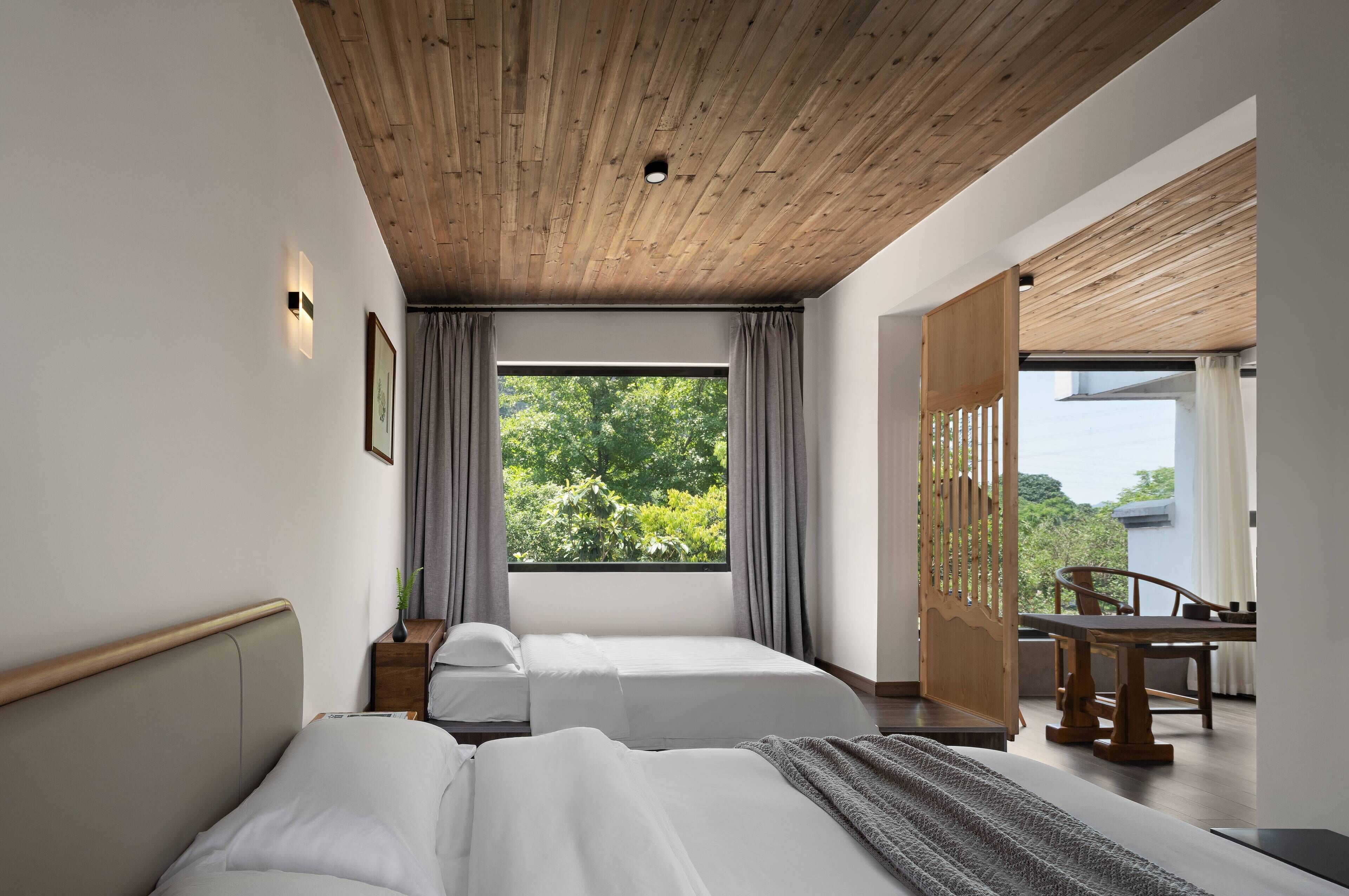 A modern hotel room with a wooden ceiling, two beds, a large window with curtains, a dining area with a table and chairs, and a view of greenery outside.