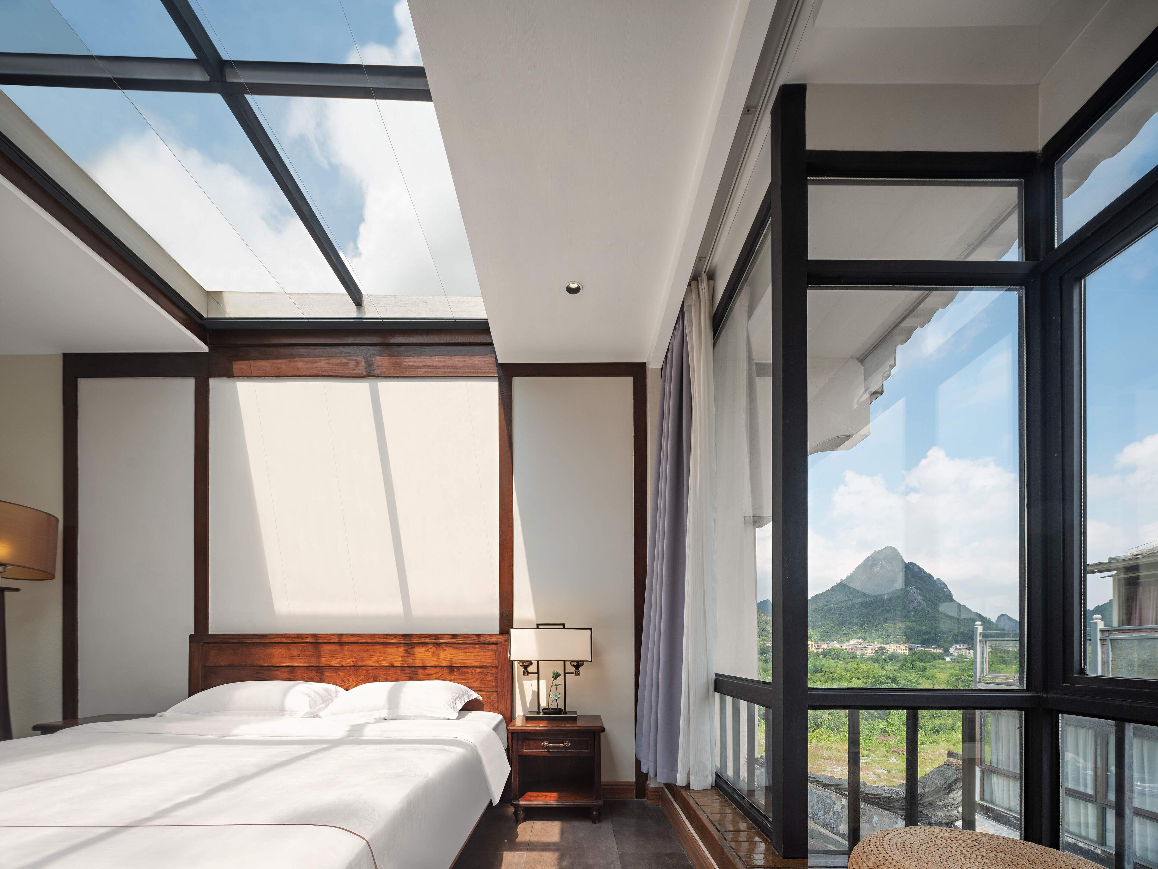 A bedroom with a large bed, a nightstand, a lamp, and a view of a mountainous landscape through large windows.