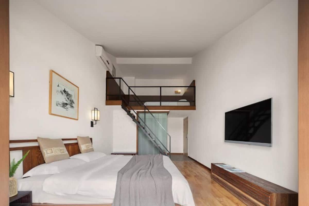 A modern hotel room with a large bed, a wooden bench, a TV, and a staircase leading to an upper level.
