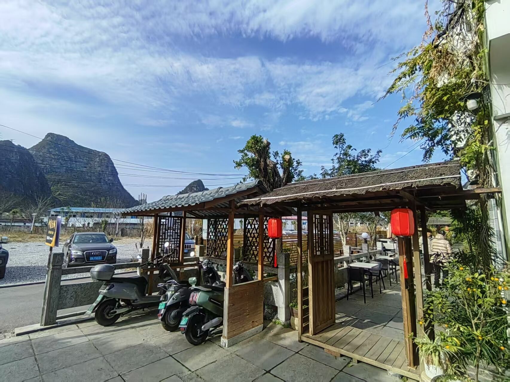 A covered outdoor seating area with a wooden structure, a car, and a mountain in the background.