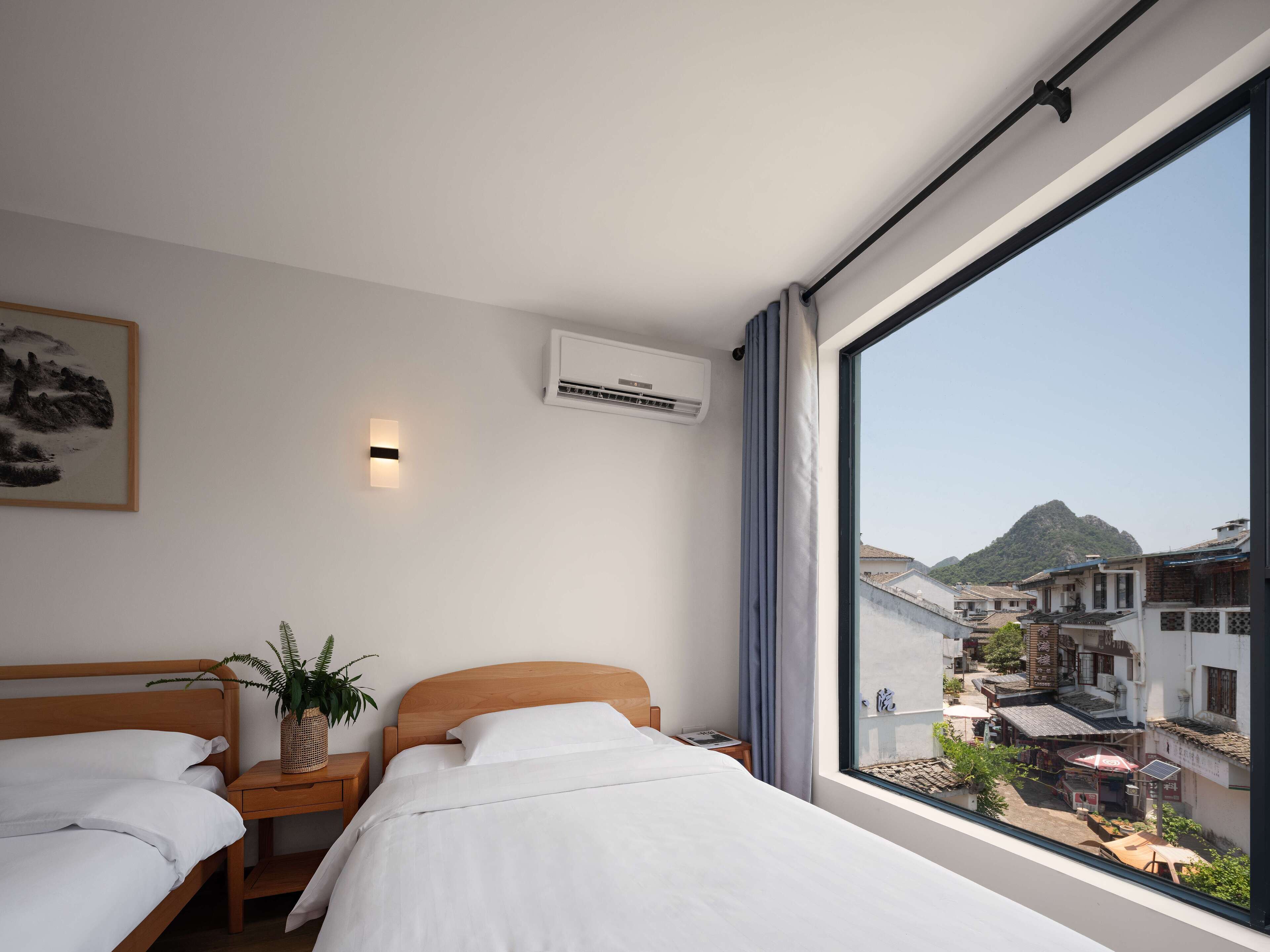 A hotel room with two beds, a large window with a view of a mountain and buildings, an air conditioning unit, and a wall-mounted light.