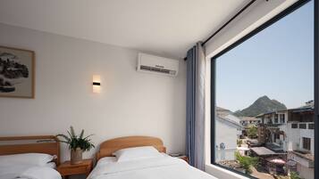 A hotel room with two beds, a large window with a view of a mountain and buildings, an air conditioning unit, and a wall-mounted light.