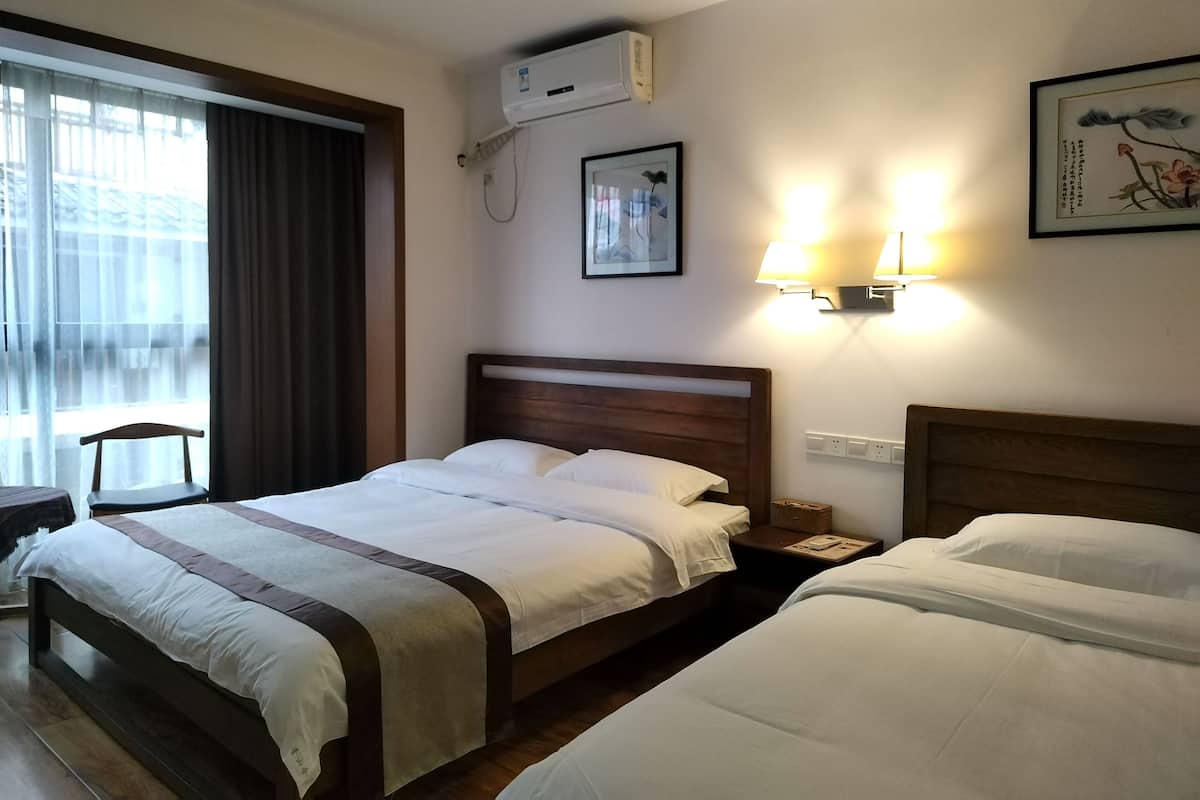 A hotel room with a large bed, a smaller bed, a desk, a chair, an air conditioning unit, and wall-mounted lamps.