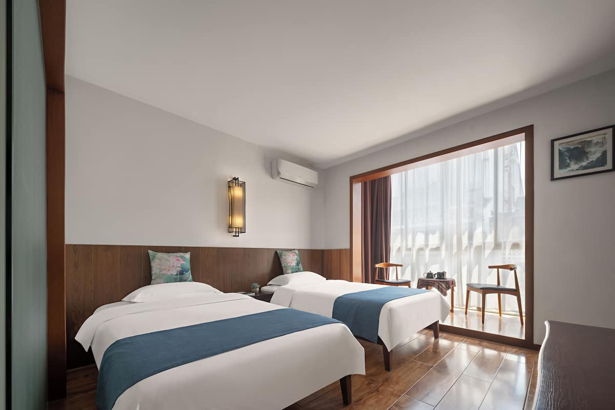 A hotel room with two beds, a large window with curtains, a wall-mounted air conditioner, and a small table with a chair.