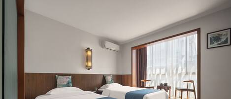 Habitación de hotel con dos camas, un ventanal con cortinas, aire acondicionado de pared y una pequeña mesa con una silla.