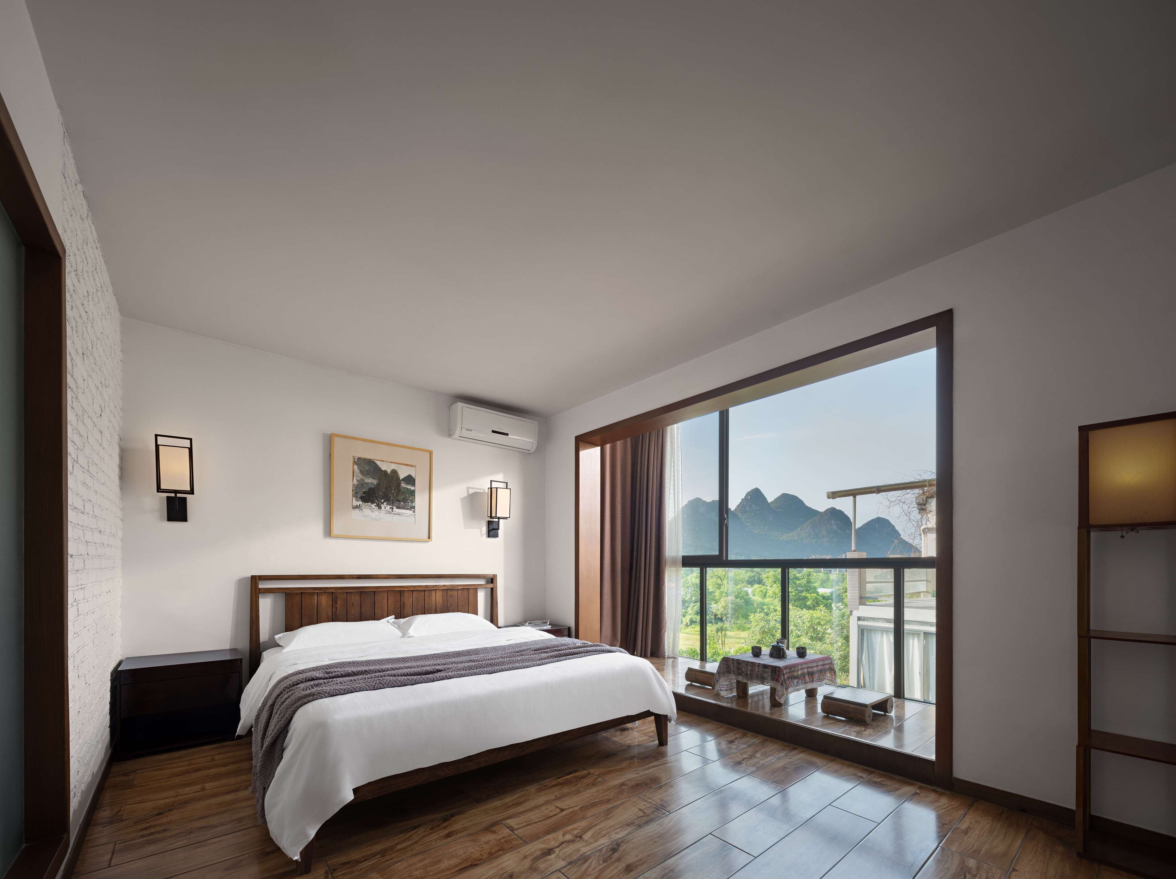 Quarto com uma grande janela, uma cama, uma mesa de cabeceira e uma pequena mesa com vista para montanhas.