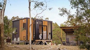 Una cabaña moderna y compacta con fachada de madera y puerta de vidrio, ubicada en un entorno natural con árboles y una estructura pequeña contigua.