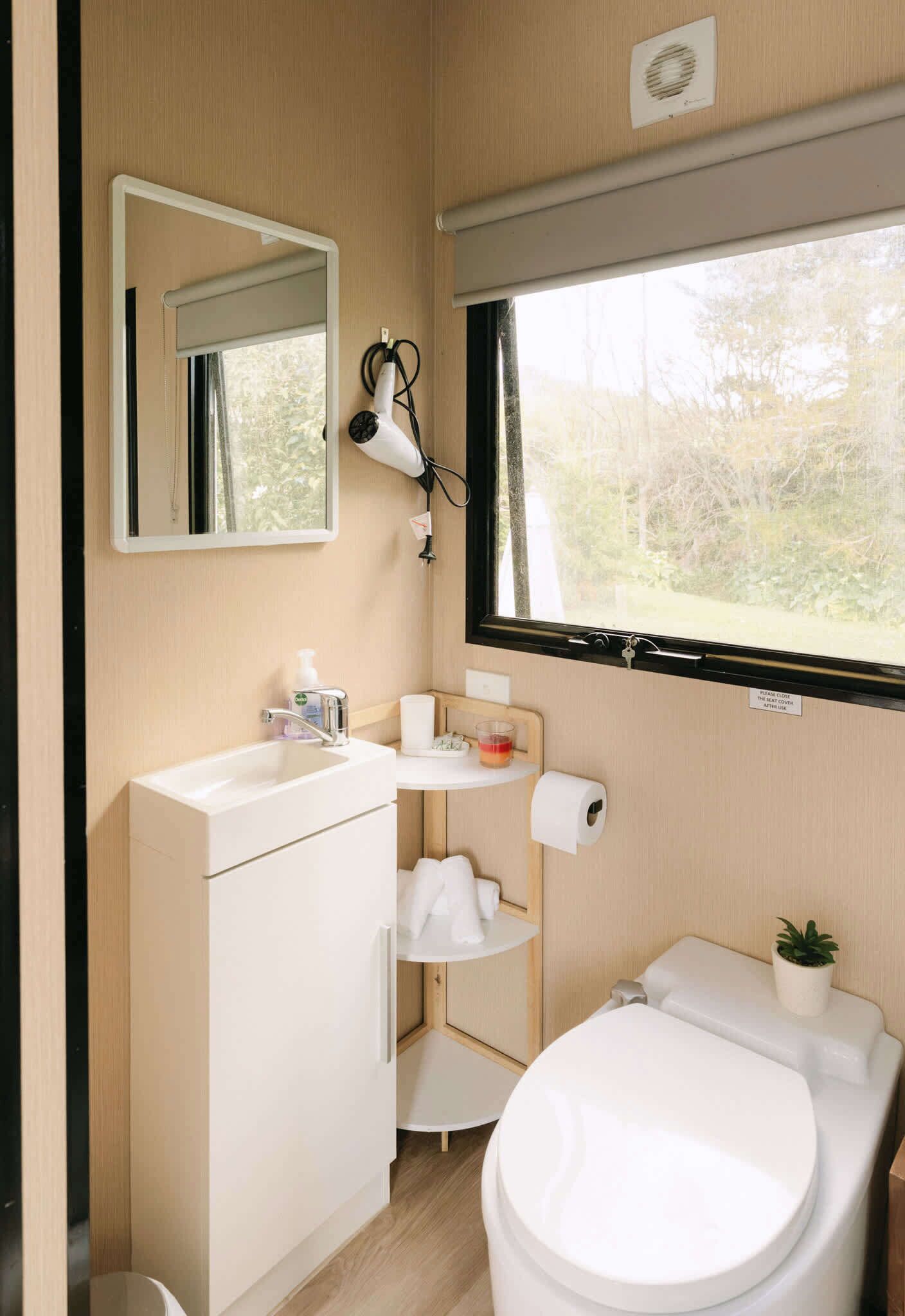 A compact bathroom with a toilet, sink, and mirror. A hairdryer is mounted on the wall, and a small shelf holds toiletries.