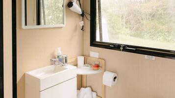A compact bathroom with a toilet, sink, and mirror. A hairdryer is mounted on the wall, and a small shelf holds toiletries.