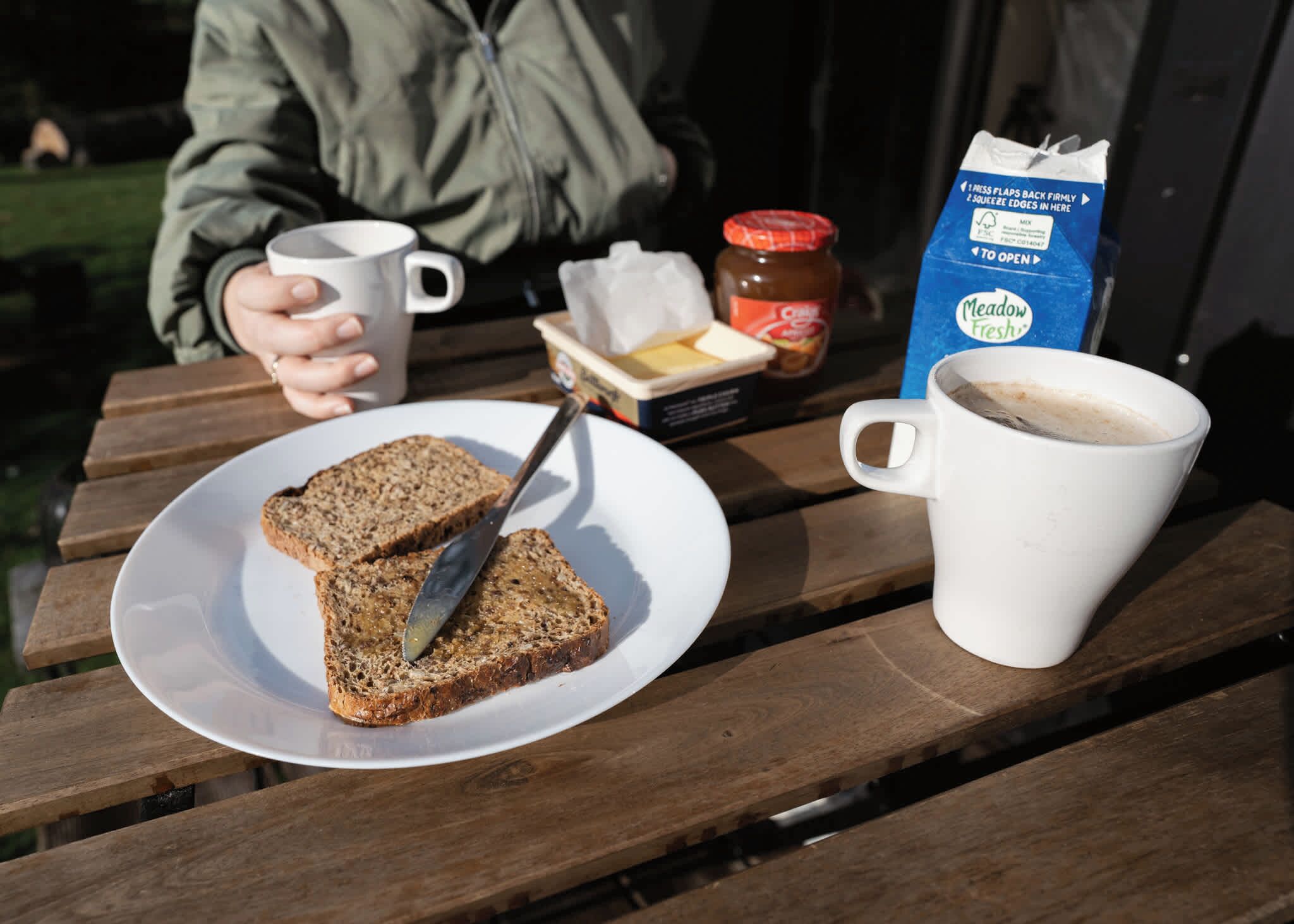 Une table en bois sur laquelle se trouvent une assiette de toasts, un pot de confiture, un paquet de beurre, une tasse de café et un carton de lait.