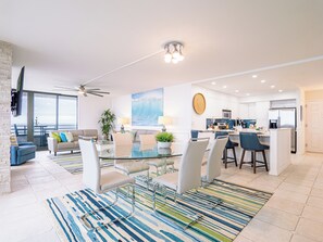 Ein modernes Wohnzimmer mit Glastisch, eine Essecke mit Barhocker, eine Küche mit weißen Schränken und ein großes Fenster mit Meerblick.