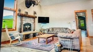 Ein gemütliches Wohnzimmer mit Kamin aus Stein, einem grauen Sofa, einem Schaukelstuhl und einem an der Wand befestigten Fernseher.