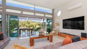 A spacious living room with a large TV, comfortable seating, a dining area with a table and chairs, and a view of greenery through large glass doors.