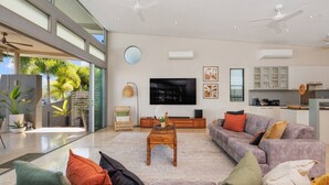 A modern living room with a large TV, comfortable seating, and a kitchen area visible through an open door.