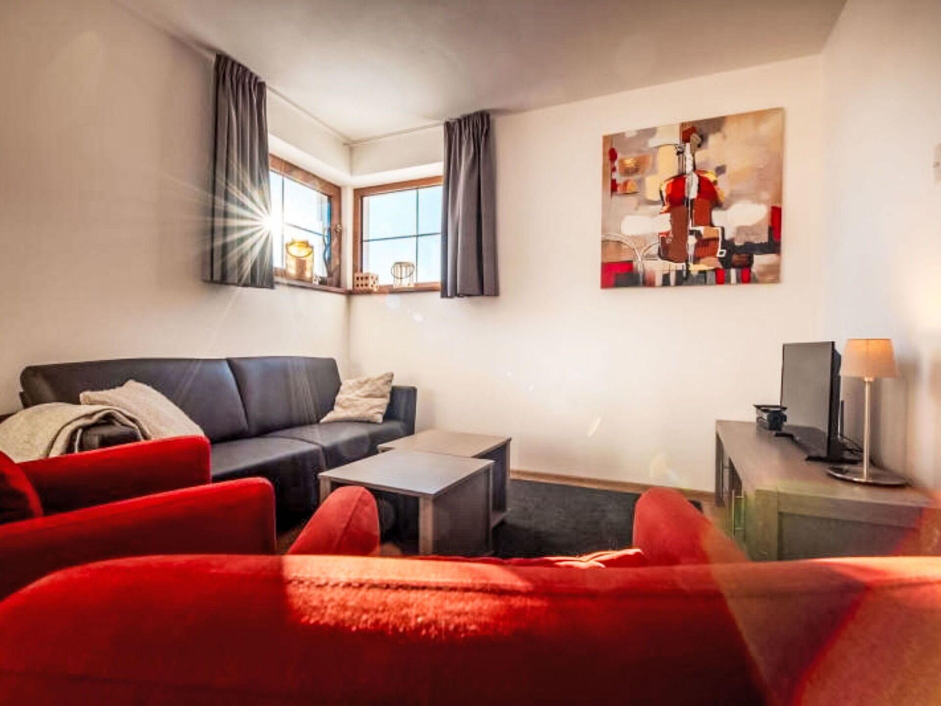 Een moderne woonkamer met een donker leren bank, een rode fauteuil, een klein bijzettafeltje en een nachtkastje met een lamp. Er is een groot raam met gordijnen en een schilderij aan de muur.